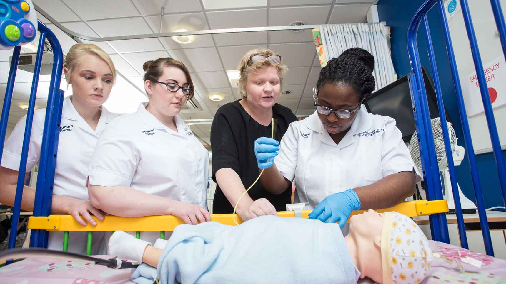 1 lecturer and 3 students stood around a hospital crib with a small simulation child being treated on. This is on the mock children's ward.