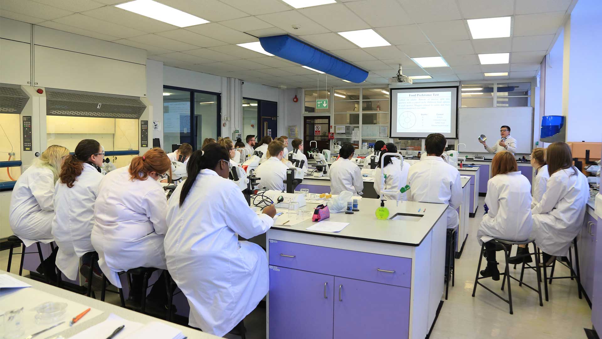 A class of students gather around the front of a science lab to listen to their teacher.